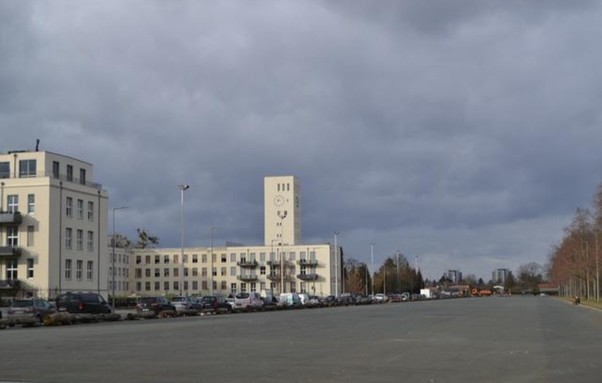Foto vom aktuellen Zustand des Platzes: Links Gebäude, dann rechts ein sehr breiter asphaltierter Streifen mit Parkplatz und grau. Am äußersten rechten Rand stehen ein paar Bäume. 