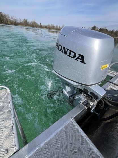 A boat's outboard motor with the "HONDA" logo in motion, creating turbulence in clear blue-green water. Diamond plate decking is visible in the foreground. Trees and open sky are in the background.