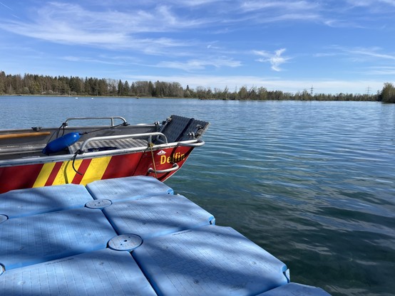 A boat with the name "Delfin" moored on a tranquil lake with blue pontoon floats in the foreground and a backdrop of trees and clear sky.