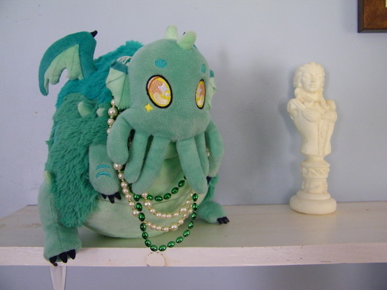A plush Cthulhu, green with wings and tentacles, wearing green and white pearl necklace, next to a statue of a woman with tentacles for hair (Lady of the Deep).