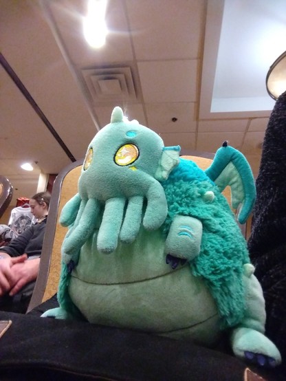 A plush Cthulhu -- green with tentacles and wings -- sitting in a chair at a conference.