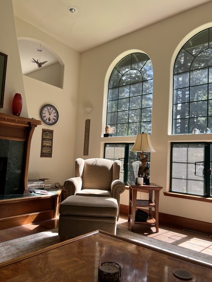 A tan chair and ottoman in a sun drenched corner of a living room, fireplace mantle and arched windows