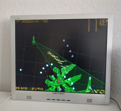 a glowing green boss made up of distinguishable parts and a pink core in it's center. it's on a black background in the lower right of the screen. also in glowing green on the upper left there's a little ship wit a cone pointing towards the boss a flurry of golden projectiles travelling in direction of the boss.