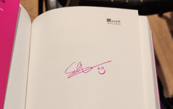 Mein Semsrott Buch mit Autogrammstempel. Man kann ein S und Schnörkel erkennen und einen Smiley. Alles in Pink.