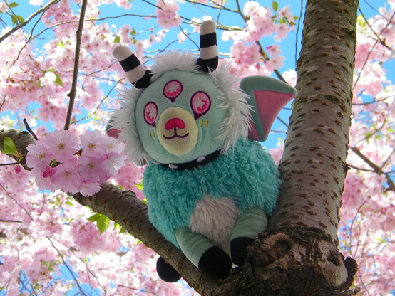 A plushie with light blue-green and white fur, black and white striped horns, three pink eyes, and pink and blue-green wings, sitting in a tree with pink blossoms. In the background is a clear, blue sky.