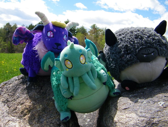 Three plush toys (one is a purple sheep, another is a green tentacled creature, and the third is a grey bat) sitting on a stone fence. Behind them is a green field and a forest.