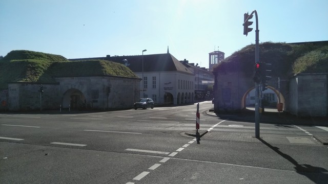 Foto einer Straßenkreuzung am Eingang zur Altstadt von Saarlouis. Die Straße wird links und rechts von alten eingeschossigen Festungsgebäuden flankiert, deren Dächer komplett grün überwachsen sind. Es ist ein strahlendheller Morgen und nur ein Auto in Anfahrt auf die Kreuzung.