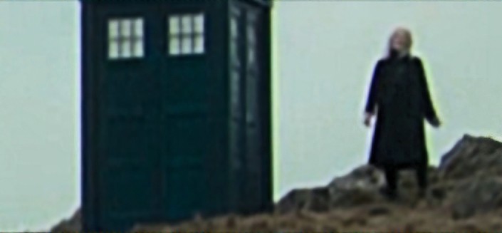 An unknown character standing on a rocky outcrop next to the TARDIS in the trailer for 73 Yards. Character has blue black overcoat and a black mask over their mouth, either blonde hair or a hat.