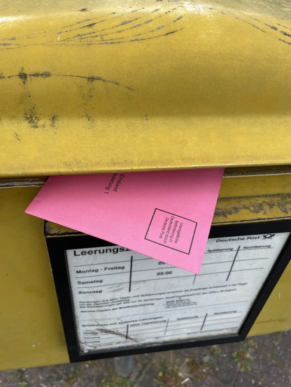 Ein roter Briefwahl-Umschlag steckt halb in einem gelben Briefkasten.