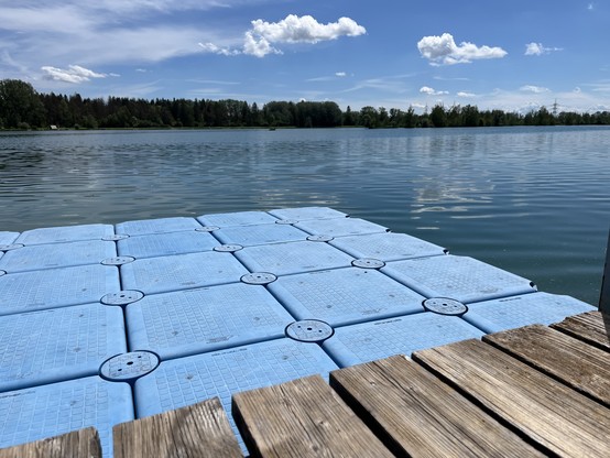 A floating blue dock on a calm lake with a forested shoreline in the distance under a blue sky with scattered clouds.
