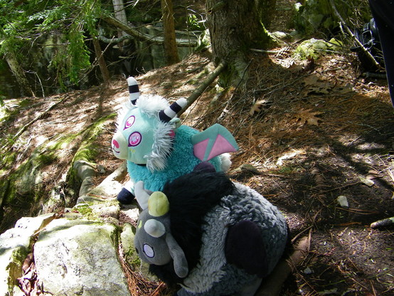 Two plush creatures sitting on mossy rocks under a tree. One is light blue-green with pink and white accents, the other is grey and black. Both creatures have horns.
