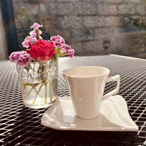 A white cup on a saucer and a glass with flowers on a table.