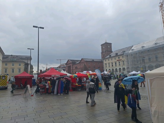 Ein Platz gefüllt mit Pavillons sowie Menschen mit mit Prideflaggen und Regenschirmen.