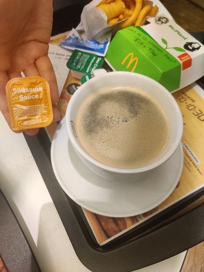 Eine große Kaffeetasse bei McDonald's, daneben eine Süßsauersauce (klein), im Hintergrund Burgerpackungen und curly fries 