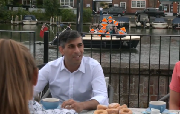 Rishi Sunak, at a campaign stop by a river, as a small boat full of Lib Dems holding party placards sails by behind him.