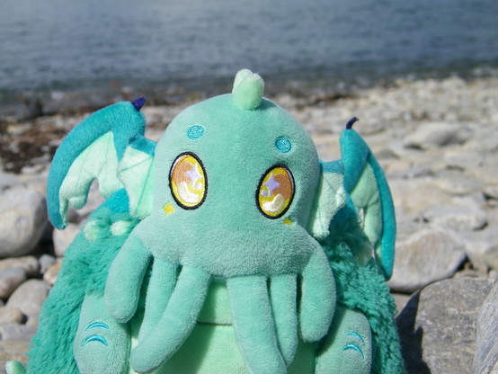 A close up of a plush creature who is light blue-green, with glowing gold eyes, tentacles, wings, and horns. Behind the plushie is a rocky shore.