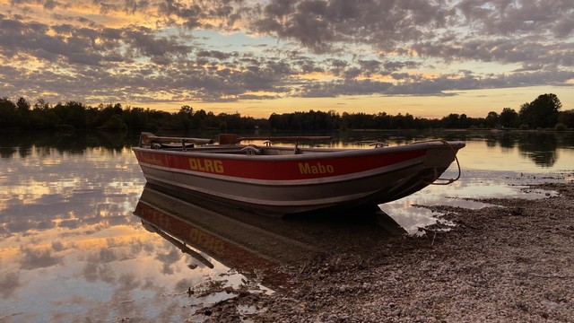 A boat labeled "DLRG Mabo" is resting on a pebbled shore at sunset, with calm water and a forested background reflecting the colorful sky.