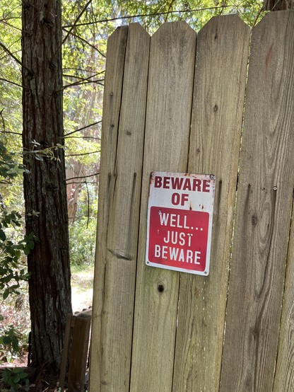 A red and white metal sign on a wooden fence in the forest “Beware of…Well Just Beware