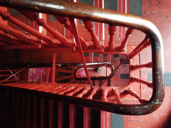 Photo von oben aufgenommen: Eine sich windende Treppe, die mehrere Rechtecke bildet. Alles ist rot, nur der Handlauf ist braun