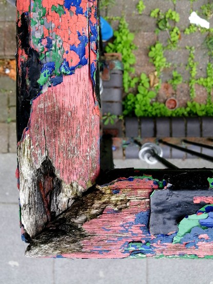 Photo von oben: ein Geländer, dessen bunter Farbanstrich abblättert, darunter sieht man rötliches vergilbtes Holz. Auf dem Boden, leicht unscharf, wächst Unkraut auf Beton.