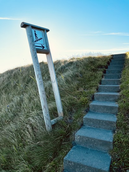 Eine Betontreppe in Dünen, daneben ein Schild an einem schiefen Pfahl mit einem Piktogramm einer Person, die Treppen steigt.