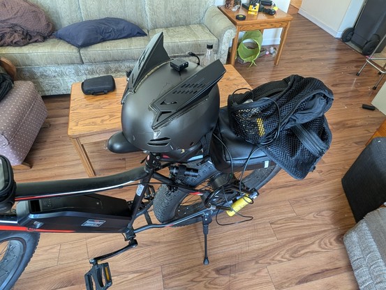 photo of my ebike in my living room, attached near the rear wheel pointing in reverse is a video camera and attached to my helmet is a front facing video camera. down the back of the helmet is a micro USB cable fed into my vest with a 6600 mAH power bank.