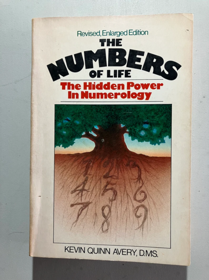 A white paperback book cover. On the bottom 2/3rds of the cover, is an illustration (in a square) of a leafy greeb tree with roots in the ground. The roots all form numbers in rows:
1 2 3
4 5 6
7 8 9

The cover type reads:

Revised, Enlarged Edition

THE
NUMBERS
OF LIFE

The Hidden Power
In Numerology

[At the bottom of the cover]
KEVIN QUINN AVERY, DMS.
