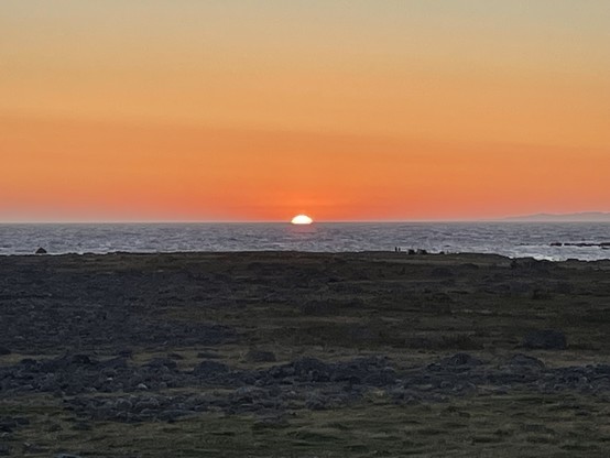 Sonnenuntergang über dem Meer. Die Sonnenscheibe ist schaut noch halb raus. Im Vordergrund steiniger Strand.