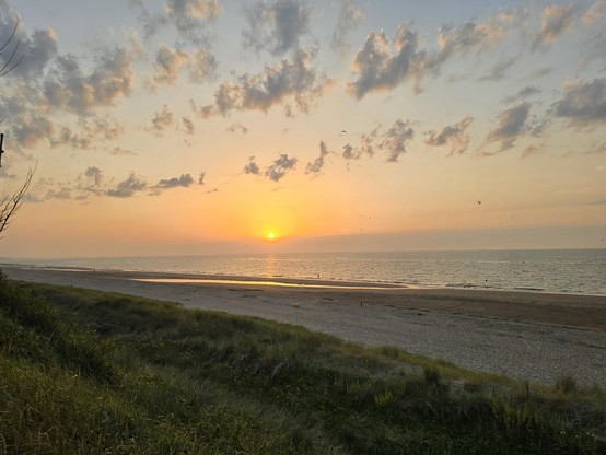 Blick über Dünen und Strand auf ein ruhiges Meer über dem die Sonne untergeht. Sie ist noch als oranger Ball über einer Wolkenbank zu sehen. Vor der Wolkenbank einzelne Schäfchenwolken.