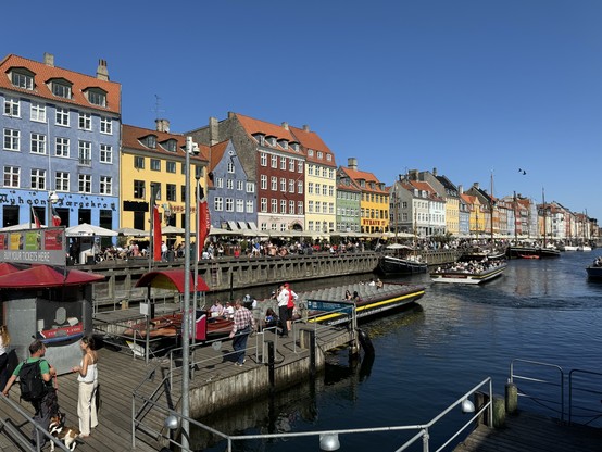 Das Bild zeigt eine malerische Hafenszene in Nyhavn, Kopenhagen. Es ist ein sonniger Tag mit klarem, blauem Himmel. Entlang des Wassers stehen bunte, historische Gebäude in verschiedenen Farben wie Blau, Gelb, Rot und Grau. Vor den Gebäuden gibt es viele Menschen, die in Cafés und Restaurants sitzen oder spazieren gehen. Im Wasser sind mehrere Boote und Ausflugsdampfer zu sehen. Auf dem Steg im Vordergrund stehen einige Personen, darunter ein Mann mit einem Hund. Die Szene wirkt lebendig und einladend.