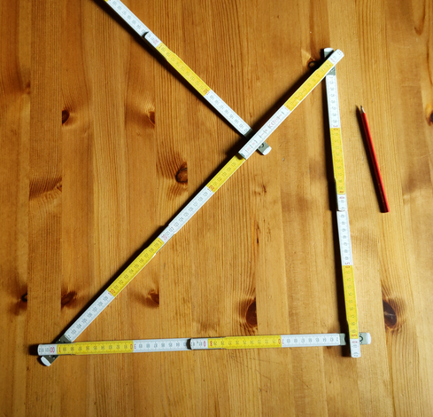 Gelb-weiß segmentierter Gliedermaßstab zu einem gleichschenkligen rechtwinkligen Dreieck geklappt, daneben ein roter Bleistift zum Anzeichnen.