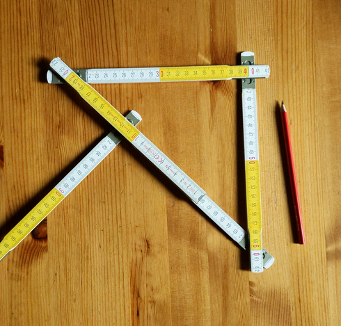 Gelb-weiß segmentierter Gliedermaßstab zu einem gleichschenkligen rechtwinkligen Dreieck geklappt, daneben ein roter Bleistift zum Anzeichnen.