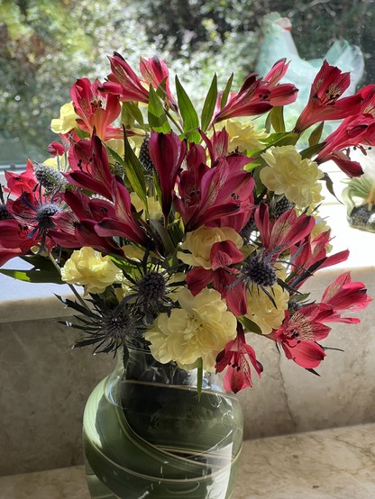 A flower arrangement, with yellow carnations, salmon colored (?) blooms, on a kitchen counter before a window 