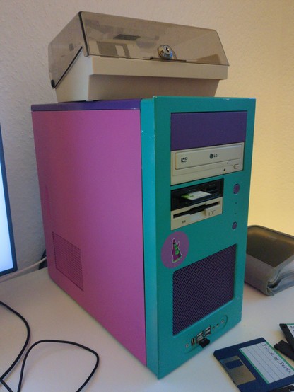 A mid sized PC tower in pink, purple and tourqouise. It has a CD/DVD drive, a (super) floppy drive, a multi card reader and a Day of the Tentacle purple tentacle sticker on the front.