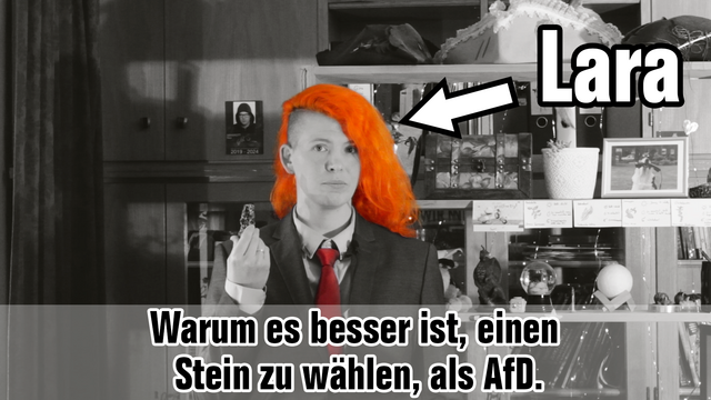 Lara (mit einem Stein in der Hand), das Bild ist bis auf die orangen Haare und die rote Krawatte komplett in Graustufen gehalten, darauf steht: 