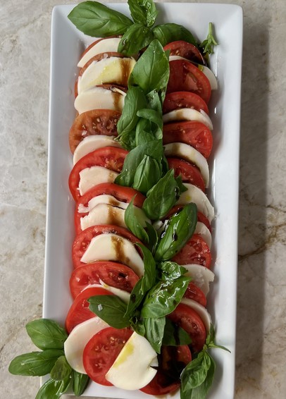 Slices of tomatoes, and slices of mozzarella with leaves of fresh basil, sprinkled with olive oil and basalmic glaze on a long white platter 