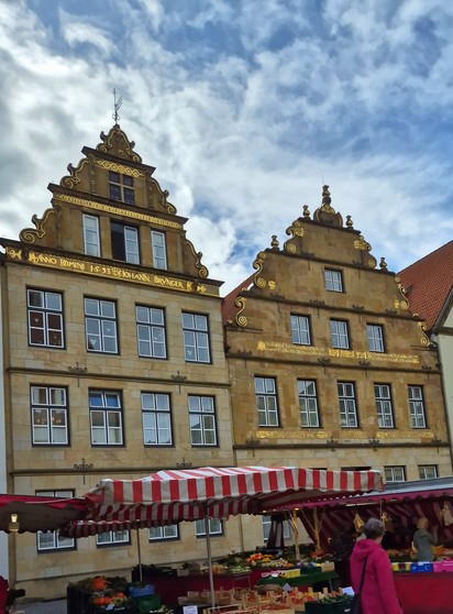 Zwei alte, mehrstöckige Häuser aus hellem Stein mit Sprossenfenstern, stufenförmigen Giebeln und goldenen Inschriften auf dem Bielefelder Markt. Vor den Häusern sind Marktstände aufgebaut.