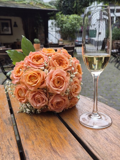 Ein Brautstrauß mit aprikotfarbenen Rosen liegt neben einem zu einem Drittel gefüllten Sektglas auf einem runden Holztisch.