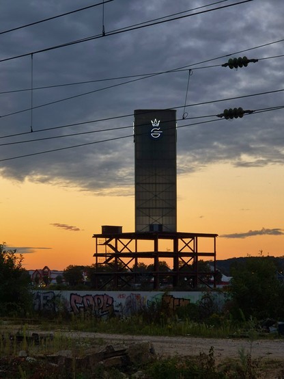 Sonnenuntergang über dem Gelände der ehemaligen Gerresheimer Glashütte in Düsseldorf. Zentral im Bild ist ein Turm mit beleuchteten G. Er steht auf einem Gebäudeskelett, was ziemlich skurril und fragil aussieht. Der Rest des Geländes ist Brache.