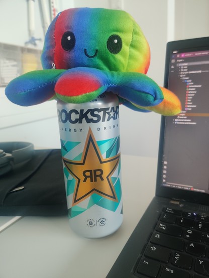 Lächelnder Regenbogenokotupus auf einem Rockstar Energy mit Laptop und IDE sowie Bank im Hintergrund 