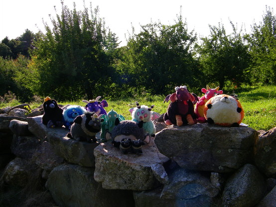 12 plushies sitting on a rock wall in an apple orchard. The plushes are a raven wearing a crown, a blue lion, a purple goat, a gray goat, a green creature with wings and tentacles (Cthulhu), a grey and white bat, two tiny grey and black goats, a brown and orange creature (Jersey Devil), a red dragon, and an orange and white fox.