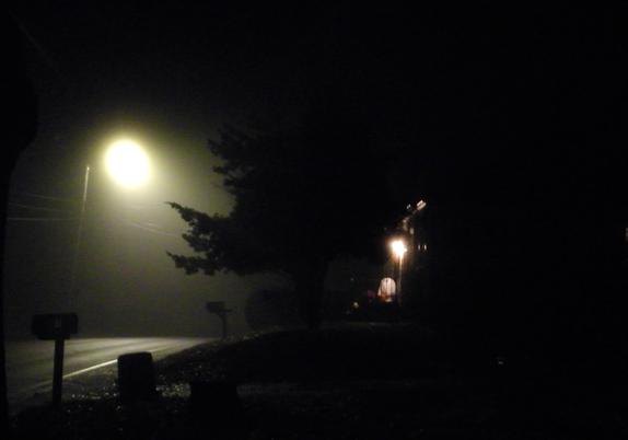 A photograph of a rural road at night. It is very foggy, and a streetlight looks like a round hazy full Moon. A fir tree is silhouetted on the side of the road. To the right of the tree is a house with a ghost-like figure and spooky lights.