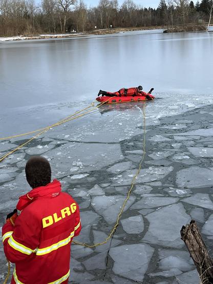 A rescue team member in a red wetsuit is lying on an inflatable rescue raft on icy water, while another team member in a red jacket with "DLRG" on the back stands on the bank, managing the rescue operation. Ice patches are visible