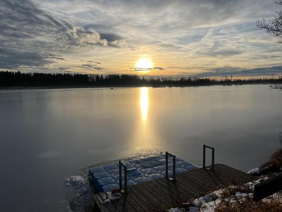 A serene lakeside view at sunset, with a wooden dock extending into smooth water reflecting the sun. The sky is filled with clouds and a warm glow, and patches of snow are visible near the water's edge.