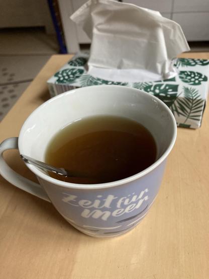 A cup of tea with a spoon rests on a wooden table. In the background, a box of tissues with a green leaf pattern is partially visible.