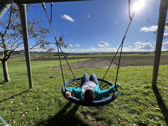 A person relaxes on a swing in a grassy area, with a scenic view of rolling hills and a blue sky filled with clouds. Sunlight shines brightly in the background, creating a peaceful outdoor atmosphere.