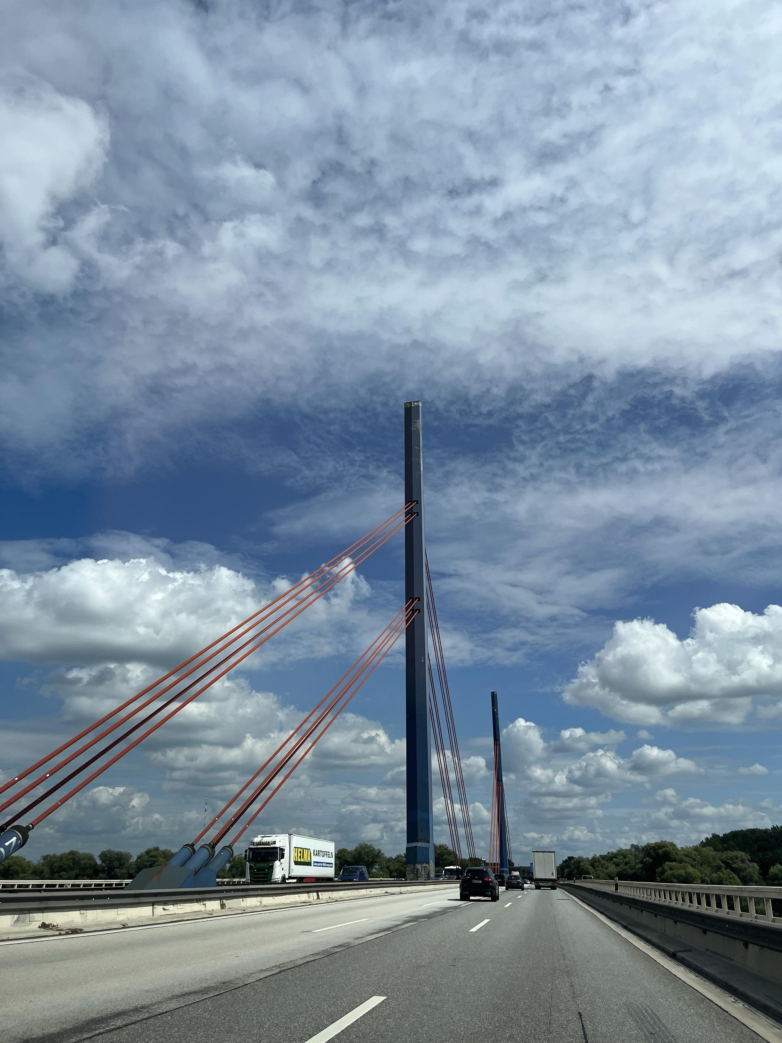A highway scene featuring a cable-stayed bridge with tall, blue towers and orange cables against a backdrop of a partly cloudy blue sky. Vehicles are traveling on the road, with a truck visible in the left lane.