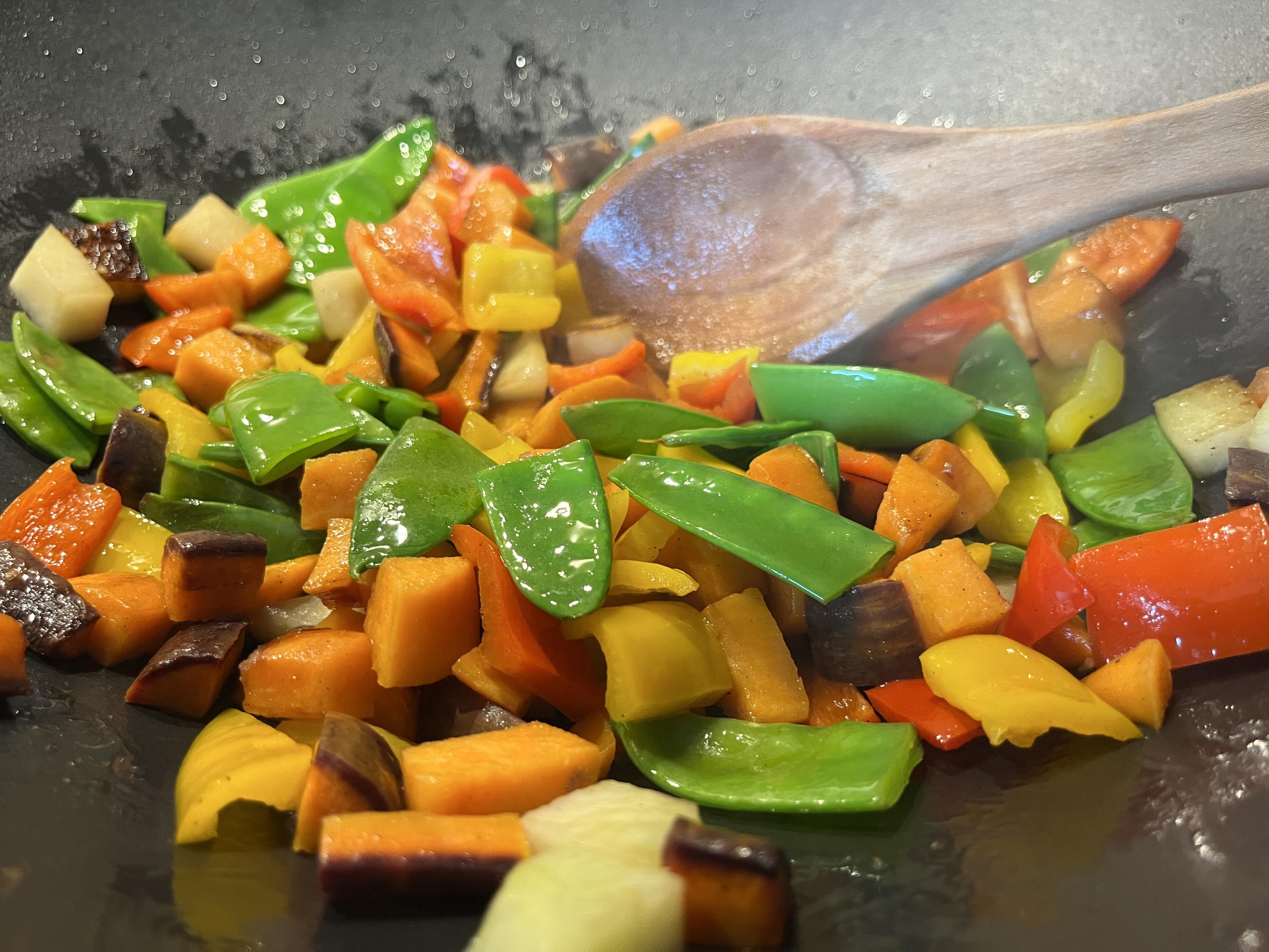 A variety of colorful vegetables, including bell peppers, snap peas, and sweet potatoes, are tossed in a pan. A wooden spoon is visible, suggesting the process of stir-frying. Steam and moisture are present, indicating that the vegetables are being cooked