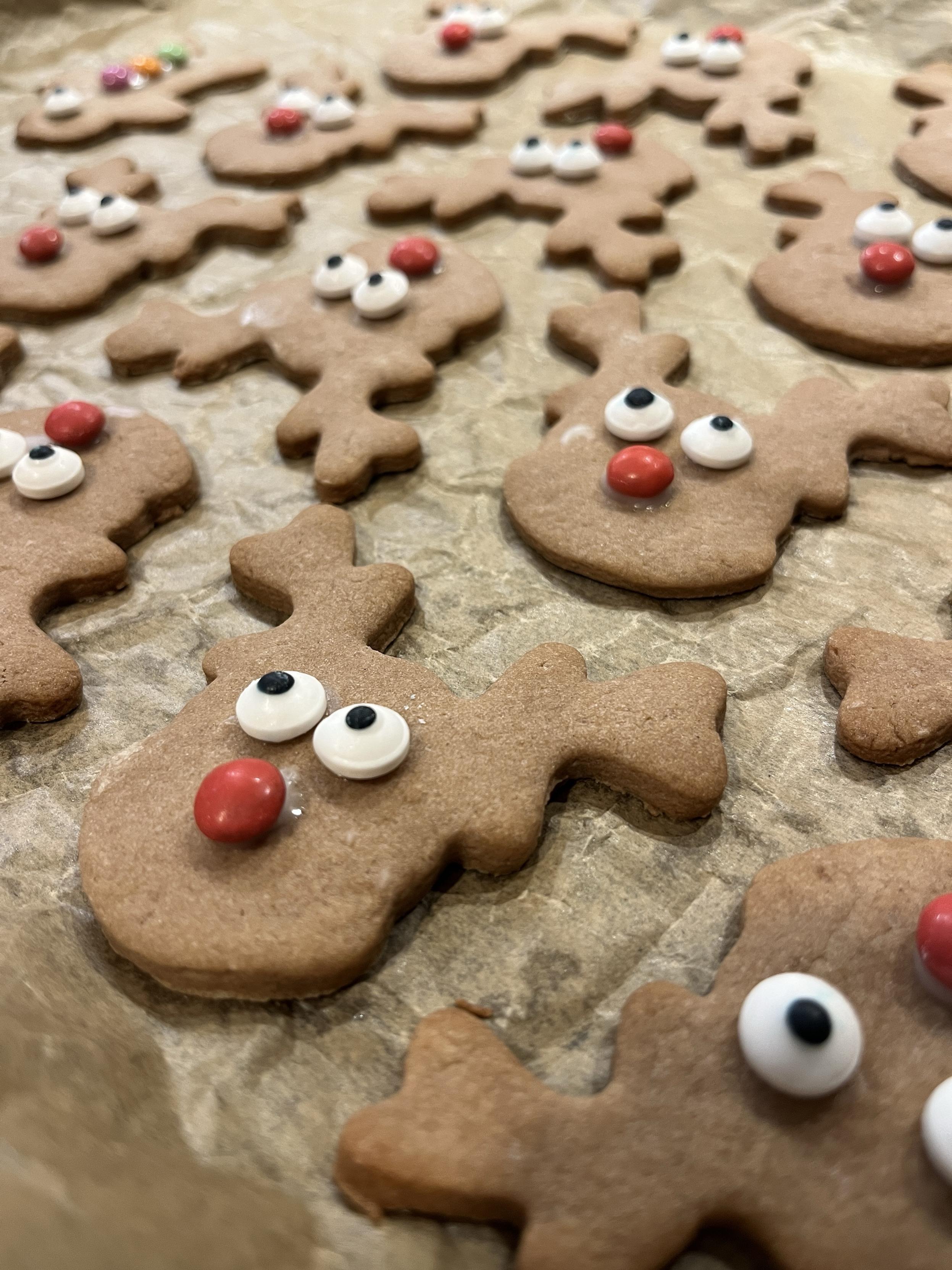 A close-up of gingerbread cookies shaped like reindeer, decorated with white and black candy eyes and red noses. They are arranged on parchment paper.